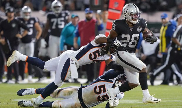 Josh Jacobs #28 of the Oakland Raiders breaks the tackle of Corey Nelson #56 and Bradley Chubb #55 ...